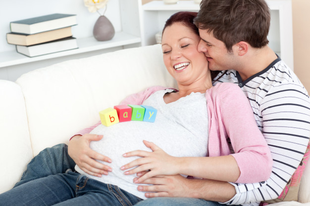 retrato-de-uma-mulher-gravida-feliz-com-cubos-de-bebe-na-barriga-e-de-seu-marido-beijando-a-em-um-sofa_13339-144848